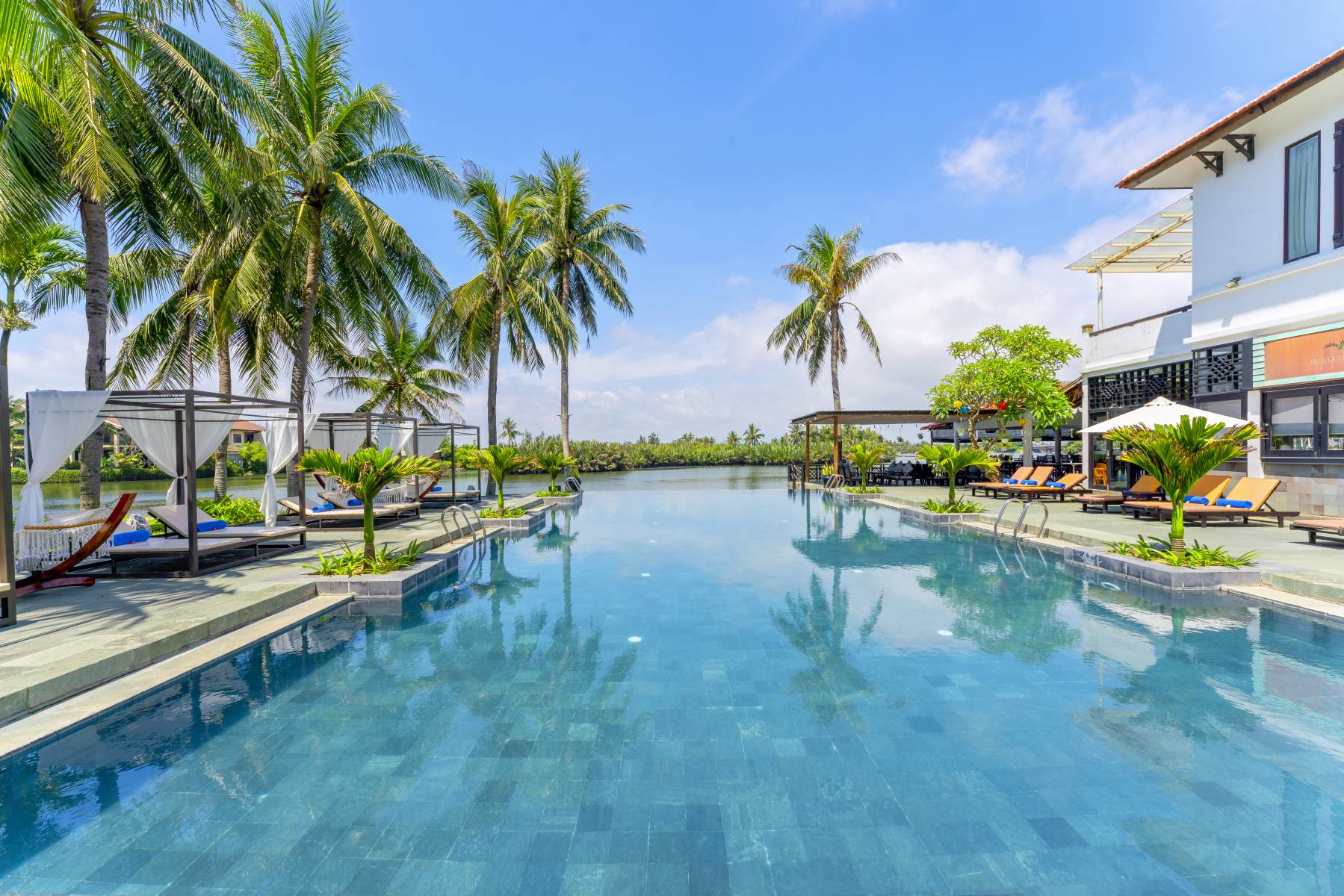 Large pool surrounded by tall palm trees and sun loungers on a bright sunny day. Your second destination, the Hoi An Beach Resort in this Vietnam holiday package
