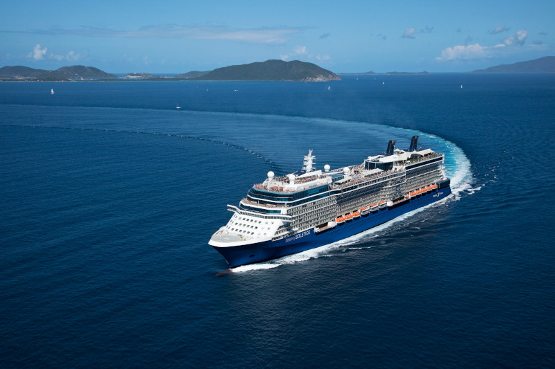 Aerial image of Celebrity Solsitce cruising in blue ocean with a lush green island in the background.