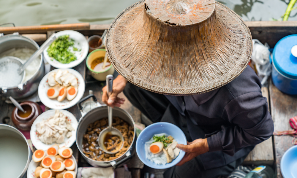 A vendor in a traditional straw hat serving food from a boat at the vibrant floating market in Thailand.