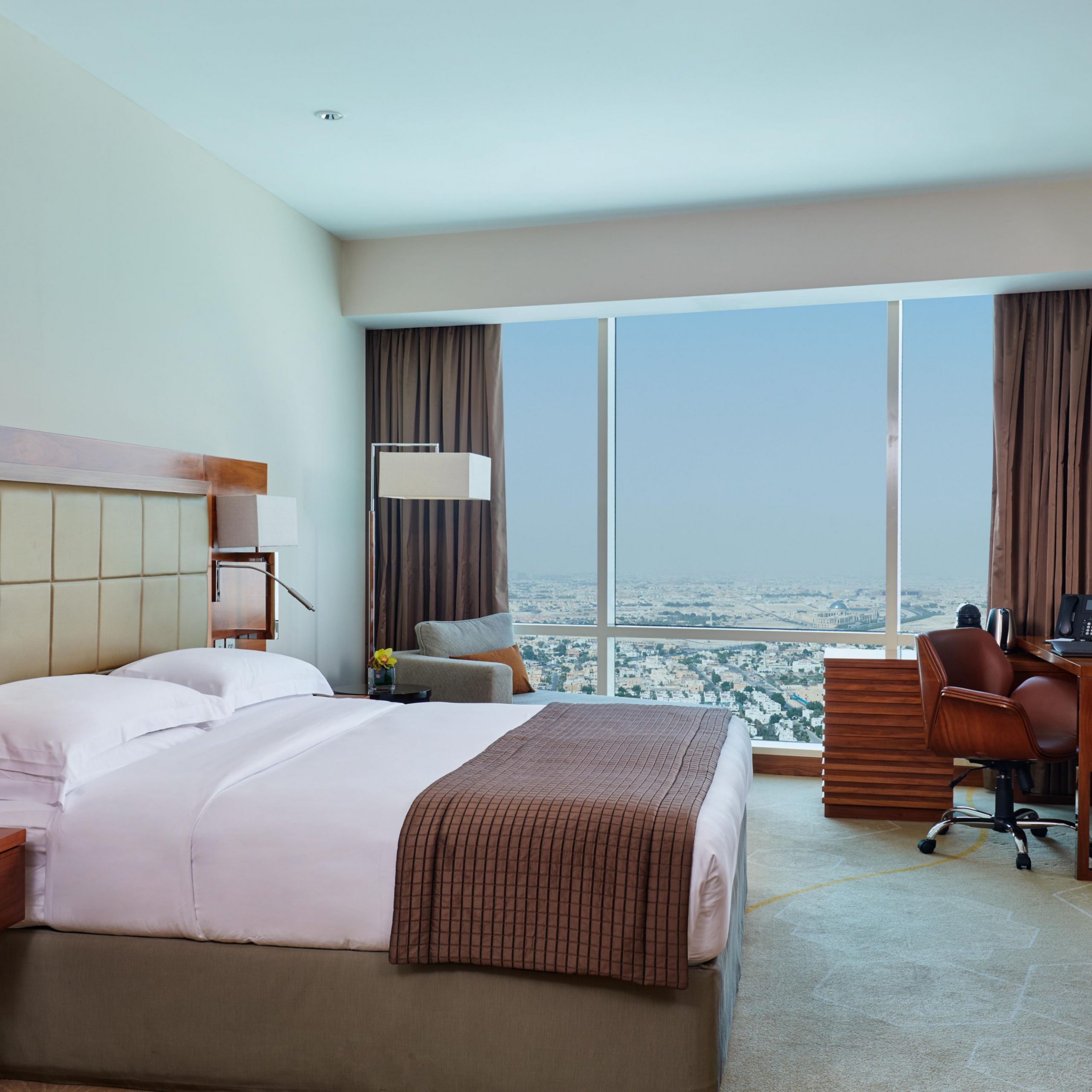 A guest suite room with bed, desk and a reclined sofa by a large window overlooking the city of Doha