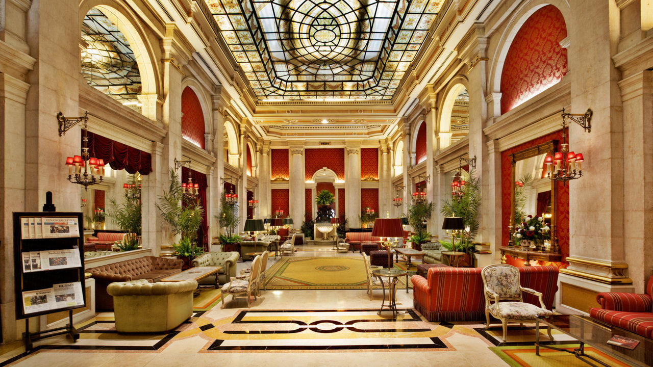 A lavish hotel lobby with red and gold accents, marble pillars, and a glass ceiling, creating a bright and sophisticated ambiance.