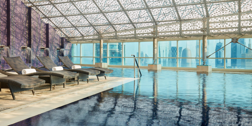 An indoor pool with loungers bathed in light with city views of Doha skyline from the windows which span its length.