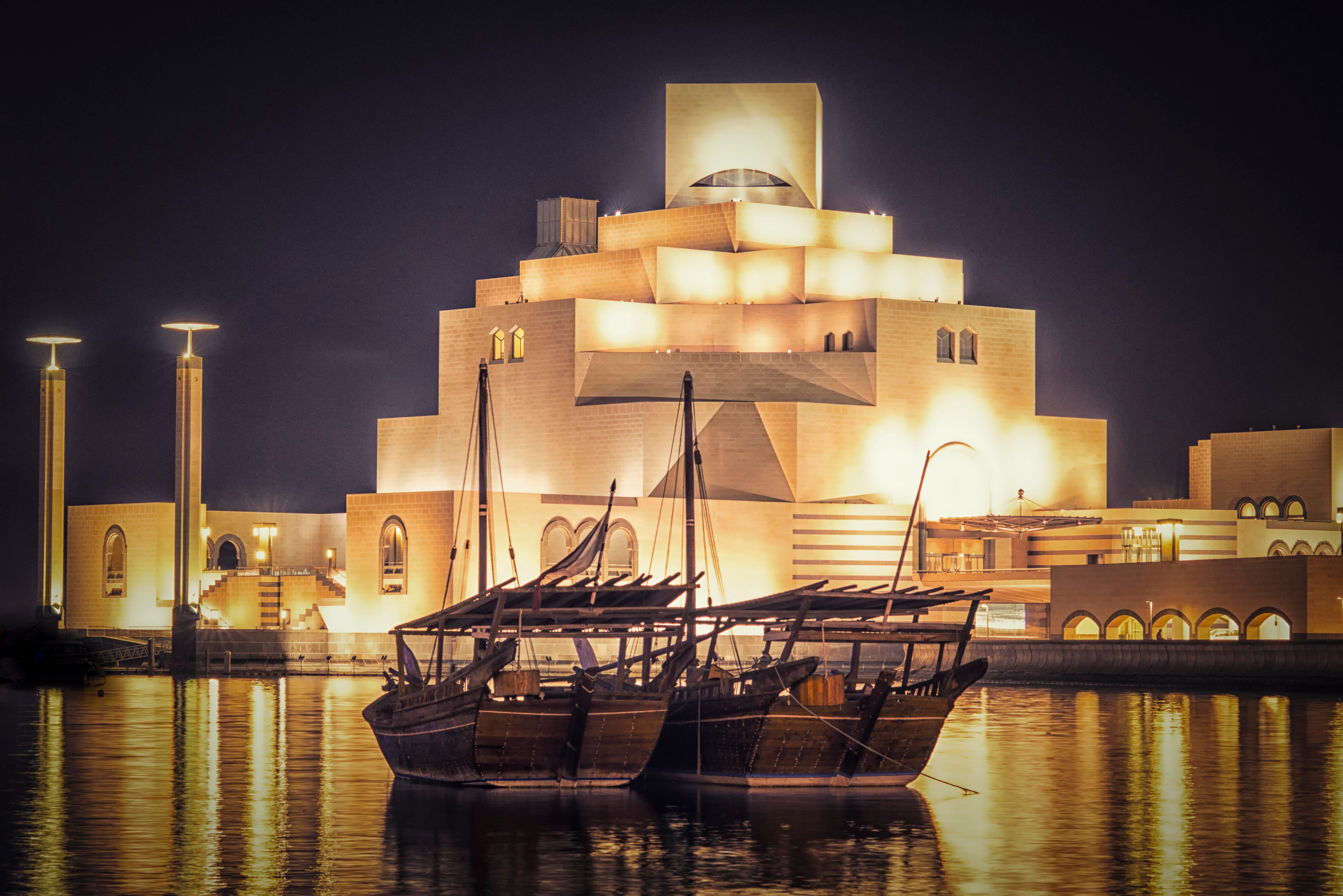 Museum of Islamic Art lit up at night with two traditional boats floating in calm water in front. Visit on our 2023 Doha Vacation Deals